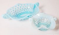 Two blue opalescent hobnail glass candy dishes with handles, one larger elongated oval and one smaller rounded square shape, shown side by side on a beige surface against a light background.