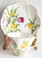 Fine bone china teacup and saucer with floral patterns of daffodils and tulips, gold trimmed scalloped edges, displayed on wooden holder.