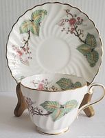 Photo of the teacup placed in front of the matching saucer showing floral pink cherry blossom branches and green leaves pattern with gold trim on the edges and handle.