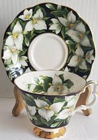 View of teacup and saucer set with floral trillium pattern on dark green background and gold trim.