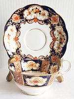 Side view of the vintage Royal Albert Crown China teacup placed in front of the matching saucer on a display stand. The cup and saucer feature detailed floral and ornate patterns in orange, gold, and navy blue on white porcelain.