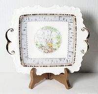 Front view of square white ceramic dish with decorative scalloped edges, central silver birch tree floral design, geometric border, and two ornate gold-tone handles.