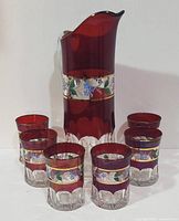 Full view of ruby flash hand painted pitcher surrounded by six matching glasses, showing color, size, and design of the set.