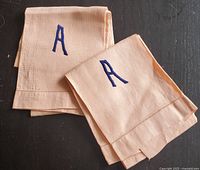 Two beige hand towels folded, showing the embroidered blue letter 'A' on each towel, placed on a dark surface.