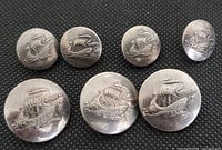 Top view of seven metal buttons grouped together on black textured surface, showing Viking ship embossing on each button.