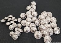 Overview of 47 vintage 1950s metal buttons with raised lion design, assorted sizes scattered on black surface.