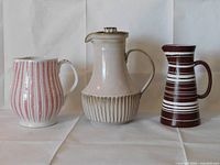 Three vintage ceramic pitchers of different styles and colors arranged side by side on white cloth background.