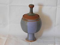 Front view of blue-gray pottery vessel with wide rounded body, pedestal base, and lid with knob. Against white background.