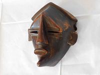 Front angle view of small African tribal wooden mask showing geometric nose, slit eyes, and carved mouth with patina on wood.