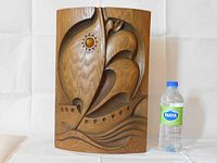 Front view of hand-carved walnut wall art depicting a stylized ship with sails and sun, with water bottle for size reference.