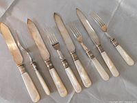 Front view showing entire 8-piece cutlery set with mother of pearl handles and metal collars, laid out on white cloth.