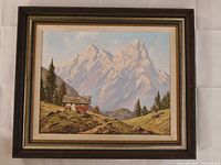 Framed oil painting showing mountain landscape with cabin, pine trees, and peaks in background, in natural tones with original period frame.
