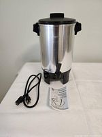 Full view of the coffee maker percolator showing stainless steel body, black lid, black base, handles, power cord and instruction manual on a white surface.