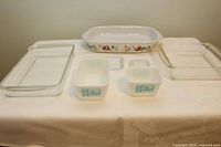 Seven baking dishes displayed on a tablecloth; includes clear rectangular dishes, Corelle fruit rim dish, two small white square dishes with blue roosters and lids