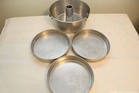 One angel food cake pan with hollow center and three round cake pans arranged on a flat white surface.