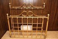 Brass headboard and footboard set shown leaning against dark wood panel wall on beige tiled floor. Both pieces polished and intact, showing the full double bed size frame components included.