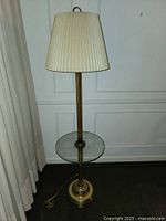 Full view of tall brass floor lamp with beige pleated lampshade and integrated round glass table.