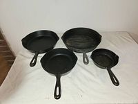 Four cast iron frying pans arranged on white cloth, including standard and grill pans.
