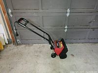 Side view of the red and black Noma TurboThrower electric snow thrower in a garage showing the handle and wheels.