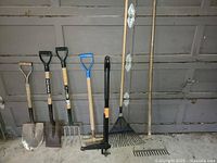 Seven garden tools lined up against garage door: two shovels, two pitchforks, a leaf rake, a garden rake, and a post hole digger.