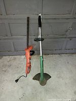 Photo showing the electric hedge trimmer and grass edge trimmer together in front of a garage door.