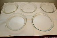 Five clear glass pie plates on white cloth, various edge designs including smooth, molded handles, and fluted edge.
