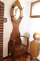 Full view of the wooden hall tree from an angle showing the oval mirror, four metal coat hooks, armchair seat with storage underneath, and decorative wood top.