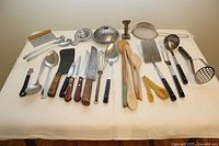 Wide shot of all kitchen utensils arranged on a white tablecloth showing knives, spoons, ladles, peelers, strainers, wooden spoons, and other tools.