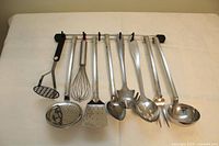 Set of 8 stainless steel kitchen utensils hanging on a metal rack including potato masher, whisk, slotted spoon, spatula, serving spoon, pasta server, fork, and soup ladle, all with metal handles and polished finish.