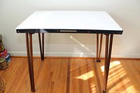 Front view of the antique metal table showing white top, black metal edge, and four tubular metal legs on a wood floor.