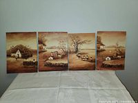 Four oil paintings aligned horizontally on white tablecloth showing interconnected rural scenes in sepia tones.