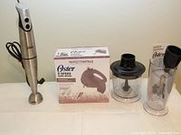 Oster hand mixer alongside its original box, food processor attachment, and whisk attachment on white surface.