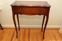 Front view of small hall table showing curved legs, drawer with metal handles, and shaped top surface.