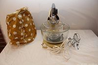 Front view of stand mixer with two glass bowls, metal beaters, power cord, and fabric cover on table