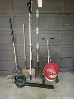 Full view of the standing garden tools including rakes, shovels, tree pruner, leaf blower on the floor, and red push spreader.