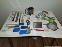 Overview photo showing all the kitchen utensils including whisks, spatulas, hand blenders, and various metal implements arranged on a white surface.