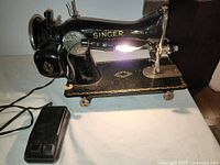Side view of the black and gold Singer sewing machine with foot pedal attached via cord, showing the sewing machine body and table base with feet.