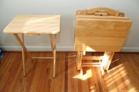 Four wooden TV trays stacked in a wooden stand on a hardwood floor with one tray opened.