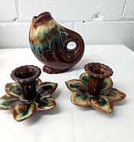 Fish-shaped ceramic pitcher with mottled brown, green, blue glaze and circular tail handle, matching flower-shaped candle holders with the same glaze on a white surface against a white brick background.