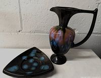 Two Blue Mountain Pottery pieces: a rainbow glaze ewer vase with a tall handle and a black triangle dish with blue glaze spots.