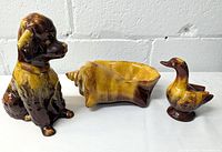 Photo showing dog figurine, shell-shaped dish, and duck figurine from a front angle on a white surface with a gray background.