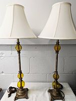 Pair of buffet lamps with metal bases and amber glass orbs, white fabric shades included. Lamps are standing on a white surface against a gray cinder block wall.