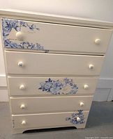 Front view of off-white dresser with five drawers, blue floral painted designs on drawer fronts, round knobs.
