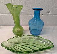Photo showing the three items: a ruffled green glass vase, a blue glass vase, and a green leaf-shaped glass dish against a white wall background.