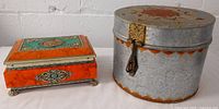 Two vintage metal tins displayed side by side on a white surface with a white brick background, front view showing details of lids and clasps
