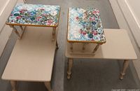 Photo showing the pair of two-tier side tables painted cream with floral decoupage on the top tier and plain cream lower tier, highlighting the detailed floral pattern and golden trim.