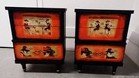 Pair of vintage nightstands showing front view with full drawer artwork and golden details on black, red and orange tone painted wood.