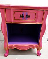Front view of the pink nightstand showing the drawer and open cubby painted purple inside with gold decorative accents on front drawer and legs.