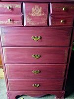 Front view of wooden highboy dresser showing 5 drawers with gold hardware and decorative gold painting on red paint.