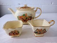 Photo showing the teapot with lid beside matching creamer and sugar dish, displaying the colorful village scenes and gold trims on a creamy ivory background.